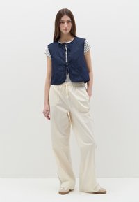 Chaleco de mezclilla acolchado de color marino, con escote redondo y cierre frontal con lazo, llevado sobre una camisa a rayas, combinado con pantalones anchos crema.