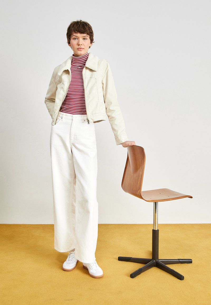 Pantalon large blanc, col roulé rayé rouge et blanc, veste courte crème, baskets blanches ; tenant une chaise en bois avec un socle en métal.