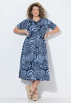 Mujer de pie con las manos en las caderas, vestida con un vestido midi azul con estampado y sandalias de cuña beige, sobre un fondo blanco.