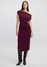Bordeauxfarbenes Off-Shoulder-Kleid mit drapiertem Stoff, tailliertem Bund mit einer goldfarbenen Schnalle und enganliegendem Schnitt, kombiniert mit kniehohen Stiefeln.