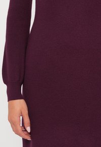 Nahaufnahme eines tiefvioletten Strickkleides, das die gerippte Textur, lange Ärmel und ein dezentes diagonales Nahtdetail zeigt.