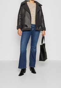 Giacca in shearling nera, maglione lavorato beige, jeans a palazzo blu, stivali neri con tacco e borsa a spalla nera. Texture morbide e colori a contrasto.