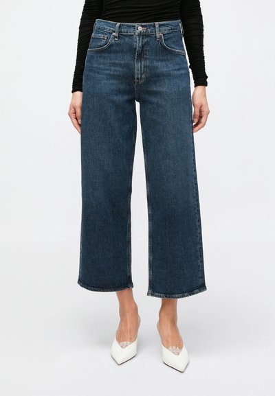 Jean large à jambe large bleu foncé avec ourlet brut, cinq poches et garnitures argentées. Le tissu a une texture lisse et une décoloration minimale.