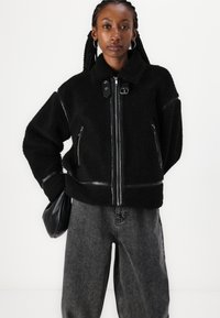 Zwarte fleecejack met leren accenten, rits aan de voorkant en zijzakken. Kenmerkt zich door een kraag en een oversized pasvorm; gecombineerd met grijze denim broeken.