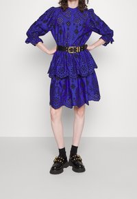 Robe violette avec broderie florale découpée, volants superposés et ourlet à volants. Associée à une ceinture noire et des chaussures noires chunky avec des accents dorés.