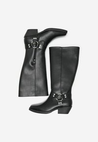 Bottes noires en cuir montantes avec un bout arrondi, un petit talon, des détails de fermeture éclair, et une sangle décorative avec des accessoires en métal argenté.