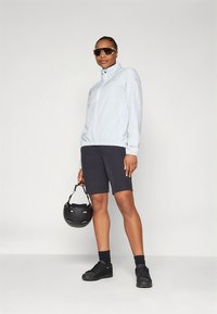 Jasnoniebieska kurtka na zamek, czarne spodenki, czarne trampki i okulary przeciwsłoneczne. Trzyma czarny kask, o gładkiej teksturze tkaniny i sportowym kroju.