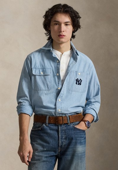 Svetlo modra denim srajca z dvema žepoma na prsih, vezavnim logotipom, oblečena preko bele henley majice, kombinirana z izbleglimi modrimi kavbojkami in rjavim pasom.