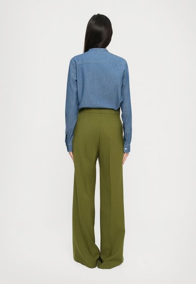 Donna con lunghi capelli scuri che indossa una camicia di denim blu infilata in pantaloni larghi a vita alta di colore verde oliva, in piedi di spalle.