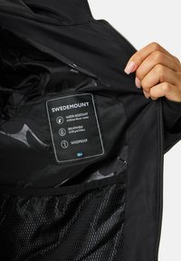 Wnętrze czarnej kurtki Swedemount z widoczną metką pielęgnacyjną. Cechy produktu obejmują materiały wodoodporne i oddychające, projekt wiatroodporny oraz siateczkową podszewkę.