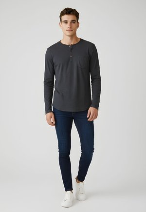 Joven de pie, vistiendo una camiseta henley de manga larga gris oscuro, jeans ajustados azul oscuro y zapatillas blancas sobre un fondo claro sencillo.