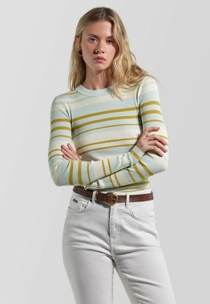 Giovane donna con capelli biondi lunghi indossa un maglione a coste bianco e a strisce giallo senape e jeans bianchi a vita alta, con le braccia incrociate.