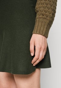 Vestido de punto con falda verde oliva y mangas marrón con textura y acanaladas. Tejido suave con un corsé ajustado y dobladillo acampanado.