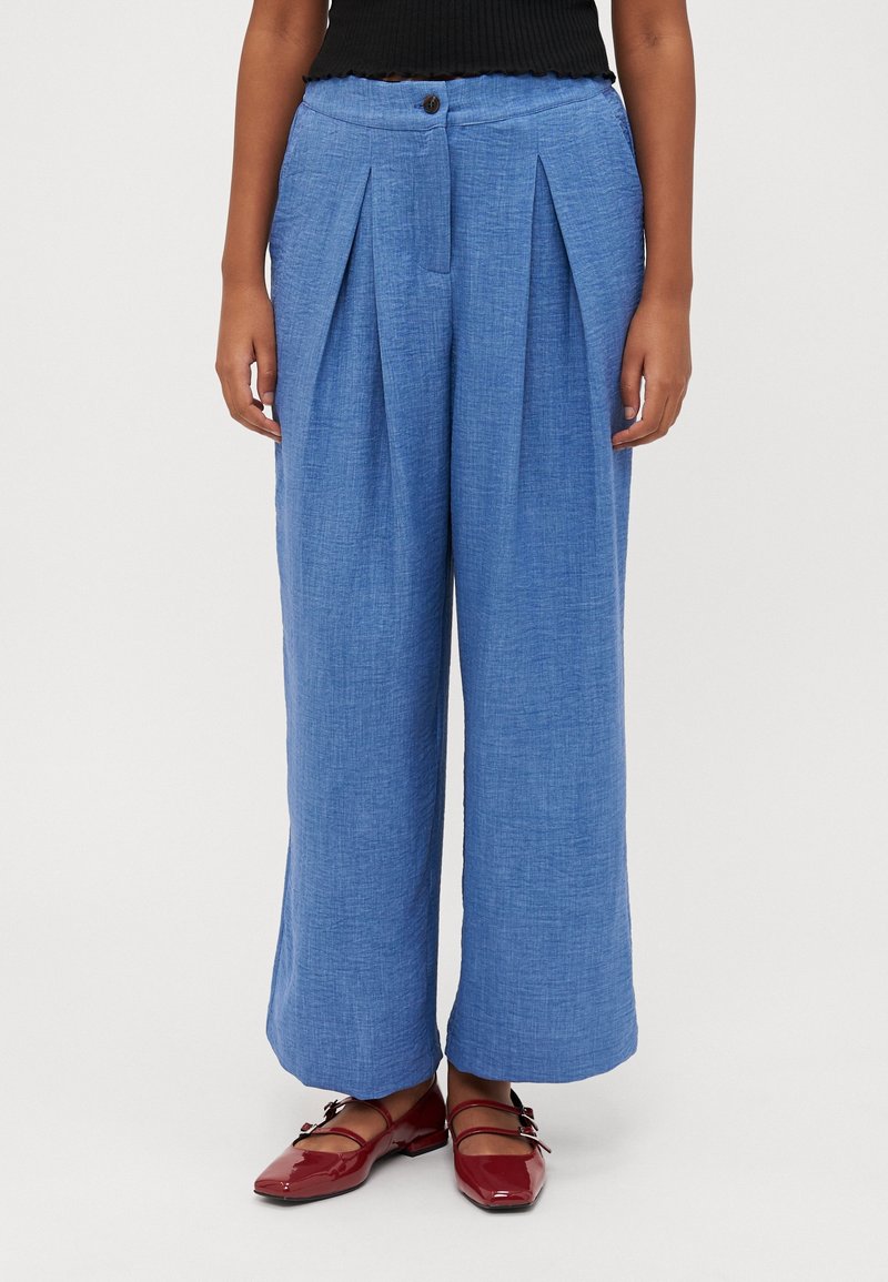Femme portant un pantalon bleu taille haute à jambes larges avec des plis devant et un haut noir, associé à des chaussures Mary Jane rouges sur fond blanc.