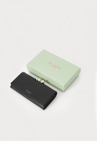 Carteira de couro preto texturizada com fecho de clasp, apresentada numa caixa verde-claro com texto vermelho. Apresenta duas aplicações metálicas no clasp.