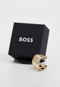 Gullfarget metallring med en bølgete design, plassert ved siden av en svart boks med merkevarenavnet "BOSS" skrevet med fete hvite bokstaver.