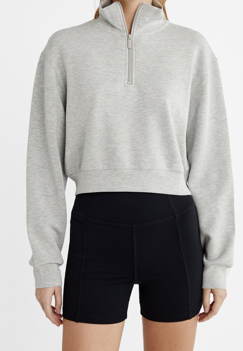 Grå, kort sweatshirt med hög krage och framzippert, kombinerad med svarta högmidjade shorts med texturerad tyg och sömnadsdetaljer.