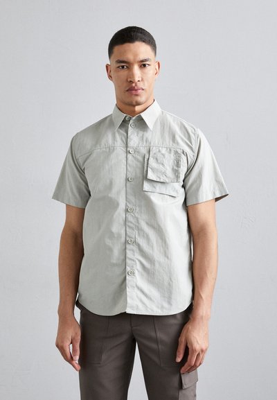 Short-sleeve, light grey button-up shirt with a chest pocket, featuring a smooth texture and a curved hem. No patterns or additional accents.