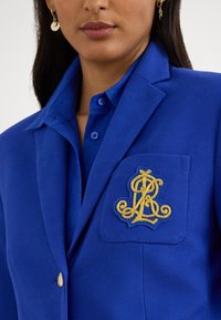 Blå blazer med teksturert vev; har en gyllen brodert logo på lommen og en kontrastfarget skjorte med knapper under.