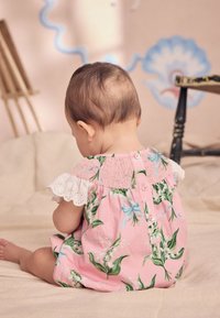 Rózsaszín virágmintás baba overál fehér csipke szegéllyel, fodros felsőrésszel és gombos záródással, zöld levelekkel és kék díszítésekkel.
