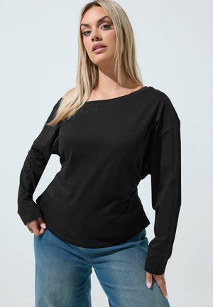 Blonde Frau trägt ein schwarzes schulterfreies Langarm-Top und blaue Jeans, posiert mit einer Hand in der Hüfte und neutralem Gesichtsausdruck.
