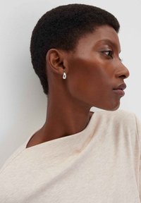 Orecchini a goccia d'argento, superficie liscia, design minimalista. Il modello indossa un top beige chiaro, strutturato, con scollo a funnel.