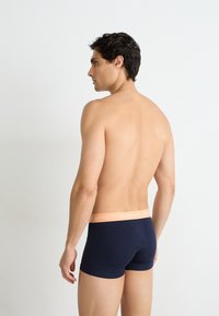 Dunkelblaue Boxershorts aus Baumwolle mit einem kontrastierenden hellorangenen Bund, auf dem der Markenname zu sehen ist. Modell steht mit dem Rücken zur Kamera.