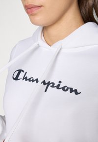 Hoodie blanc en coton avec une texture douce, présentant un logo "Champion" noir sur la poitrine et des cordons pour la capuche.