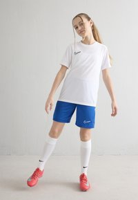 Nuori tyttö valkoisessa urheilupaidassa ja sinisissä shortseissa, valkoiset polven yli ulottuvat sukat ja punaiset jalkapallokengät, seisomassa yksinkertaisen seinän edessä.