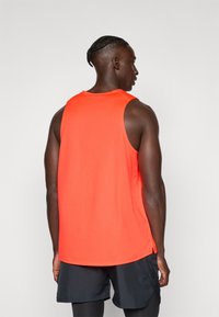 Mann med flettet hår som har på seg en lys oransje ermeløs treningsskjorte og svarte shorts, vendt bort fra kameraet mot en enkel bakgrunn.