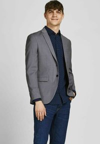 Grå jakkesætjakke parret med en marineblå skjorte med prikker og blå jeans. Jakken har en skræddersyet pasform, notch-lapper og knaplukninger ved manchetterne.