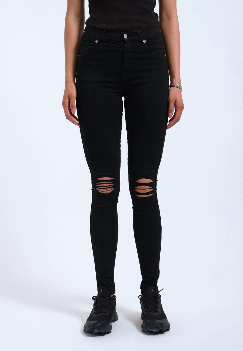 Jeans negros ajustados de cintura alta, con múltiples desgastes en las rodillas. Combinación con zapatillas deportivas negras. Textura suave.