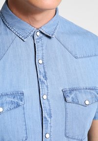 Acercamiento a una persona que lleva una camisa de mezclilla azul claro con botones de presión blancos y dos bolsillos en el pecho con solapas.