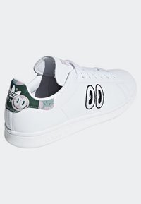 Baskets blanches avec une tige en cuir lisse, un bout rond, des yeux brodés sur les côtés et un contrefort de talon à motif vert et rose.