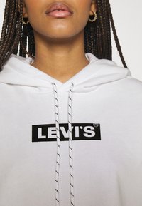 Sudadera blanca con un logo negro de "LEVI'S" en un parche rectangular, con cordones de rayas negras y blancas, textura de material suave.