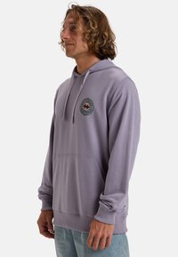 Felpa viola chiaro con tasca frontale, caratterizzata da un design del logo circolare in verde e nero sul lato sinistro del petto, realizzata in tessuto morbido.