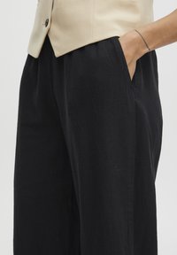 Černé kalhoty s širokými nohavicemi, vyrobené z texturovaného materiálu, s bočními kapsami a volným střihem, doplněné o béžovou přizpůsobenou vestu.