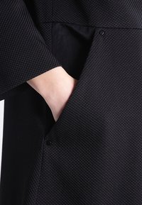 Černý texturovaný oděv zobrazující kapsu, s detailním záběrem ruky sahající dovnitř. Má tkaný vzor a nenápadné patentní zapínání.