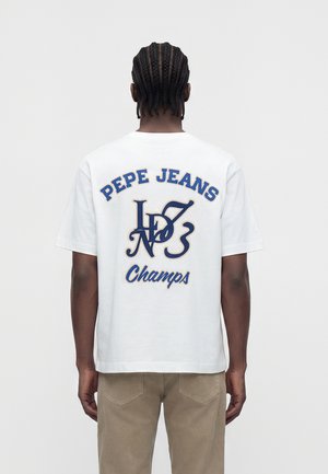 Άνδρας με πλεξούδες φοράει λευκό t-shirt Pepe Jeans με μπλε κείμενο και γραφικά, και μπεζ παντελόνι, εμφανιζόμενος από πίσω σε απλό φόντο.