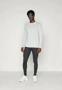 Ljust grå långärmad träningsskjorta med en liten logotyp, kombinerad med svarta leggings och vita sneakers. Reflekterande blå solglasögon bärs.