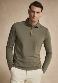 Vihreä oliivin pitkähiha polo-paita, jossa on klassinen kaulus, kolme napin aukkoa ja teksturoitu kangas, yhdistettynä vaaleisiin housuihin.
