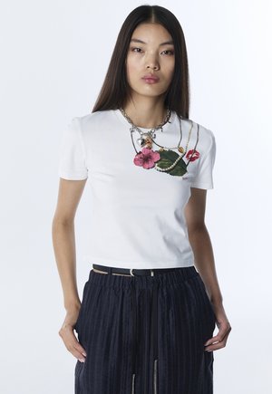 Frau mit langen dunklen Haaren trägt ein weißes T-Shirt mit Blumen-, Blatt- und Kettenmuster, kombiniert mit einem schwarzen Faltenrock und einem dünnen Gürtel.