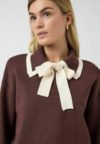 Brun genser med en kremfarget krage og sløyfedetalj. Stoffet er strikket og har en glatt tekstur, med en avslappet passform.