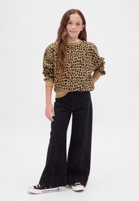 Niña con cabello largo y ondulado, vestida con sudadera de estampado de leopardo, pantalones negros de pierna ancha y zapatillas negras, sobre fondo blanco liso.
