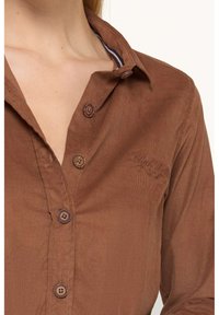 Chemise en velours c�ôtelé marron avec une conception à boutons, un col et des broderies sur la poitrine. Les boutons sont d'une teinte plus sombre.