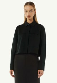 Czarna krótka bluzka z kołnierzykiem, posiadająca dwie kieszenie na piersiach i długie rękawy, wykonana z gładkiego materiału. Połączona z czarną spódnicą.