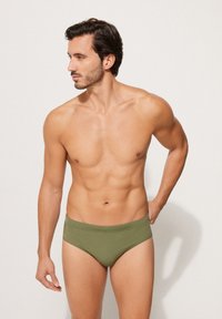 Olivově zelené plavky s jemnou, hladkou texturou, s přiléhavým střihem a elastickým pasem, předváděné mužskou postavou.