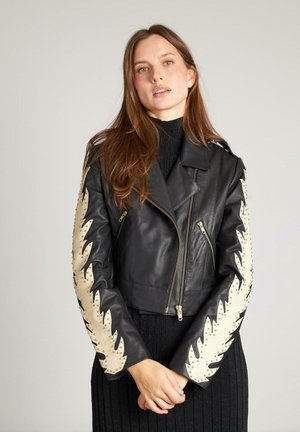 Chaqueta corta de cuero negro con detalles de patrón de hojas crema en las mangas, herrajes dorados y diseño entallado. Lleva puesta sobre un top de punto negro.