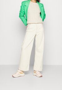 Zelené sako, béžový svetr s copánkovým vzorem, krémové kalhoty s širokými nohavicemi a světle růžové tenisky s hnědými detaily, zobrazené na jednobarevném pozadí.