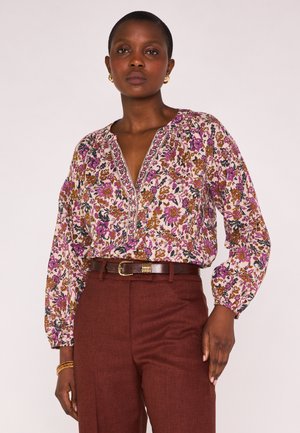 Femme aux cheveux courts portant un chemisier à manches longues à motifs floraux dans des tons violets et bruns, rentré dans un pantalon taille haute couleur rouille avec une ceinture marron.